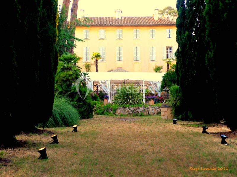 Allée bordée d'arbres menant à une grande maison jaune avec une serre et un jardin verdoyant à l'arrière-plan.
