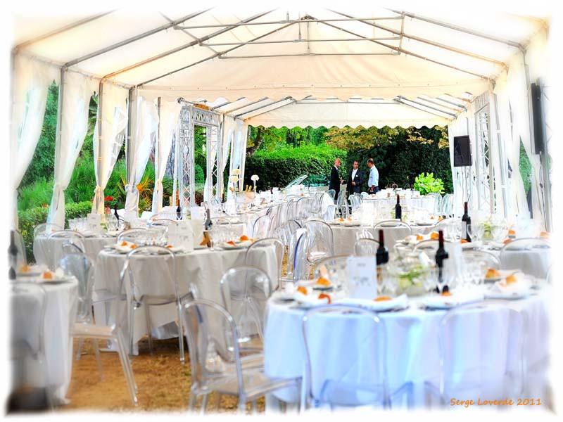 Salle de réception en extérieur sous une tente blanche, avec tables rondes dressées et chaises transparentes.