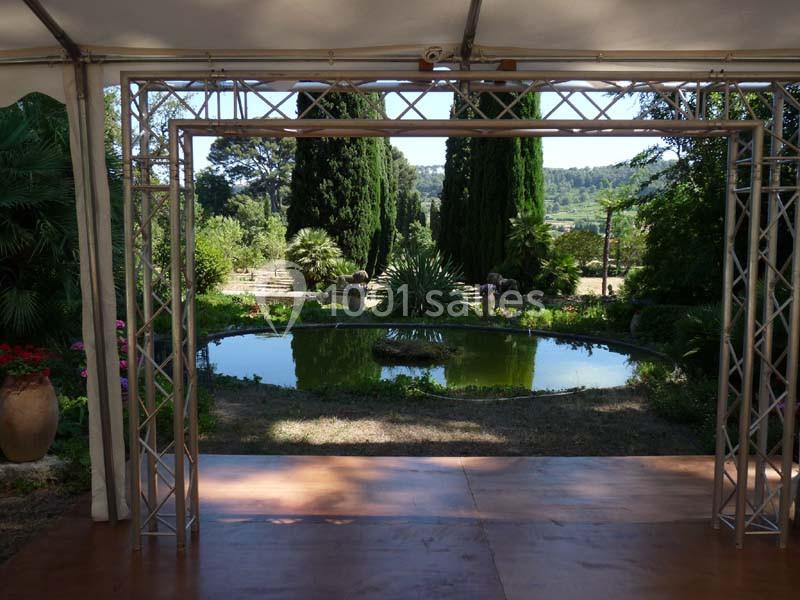 Vue d'un jardin avec un bassin rond entouré de végétation, encadré par une structure métallique sous une tente.
