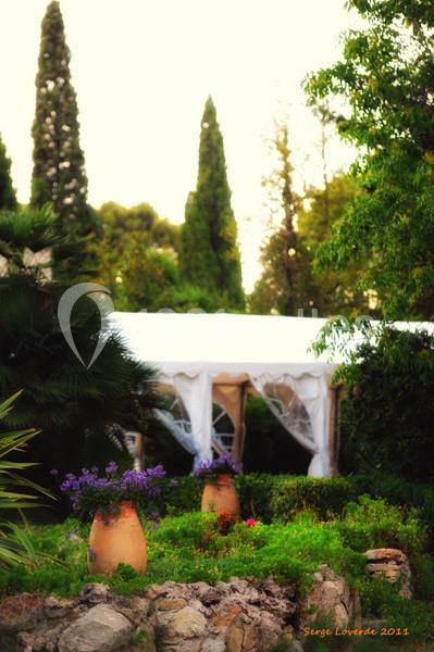 Un jardin méditerranéen avec des pots en terre cuite, des fleurs et une tente blanche entourée de verdure.