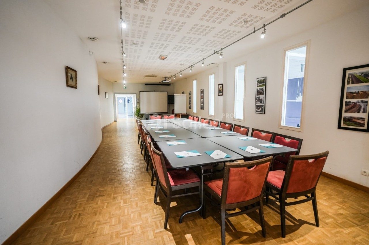 Salle de réunion lumineuse avec une grande table rectangulaire entourée de chaises rouges et un projecteur au fond.