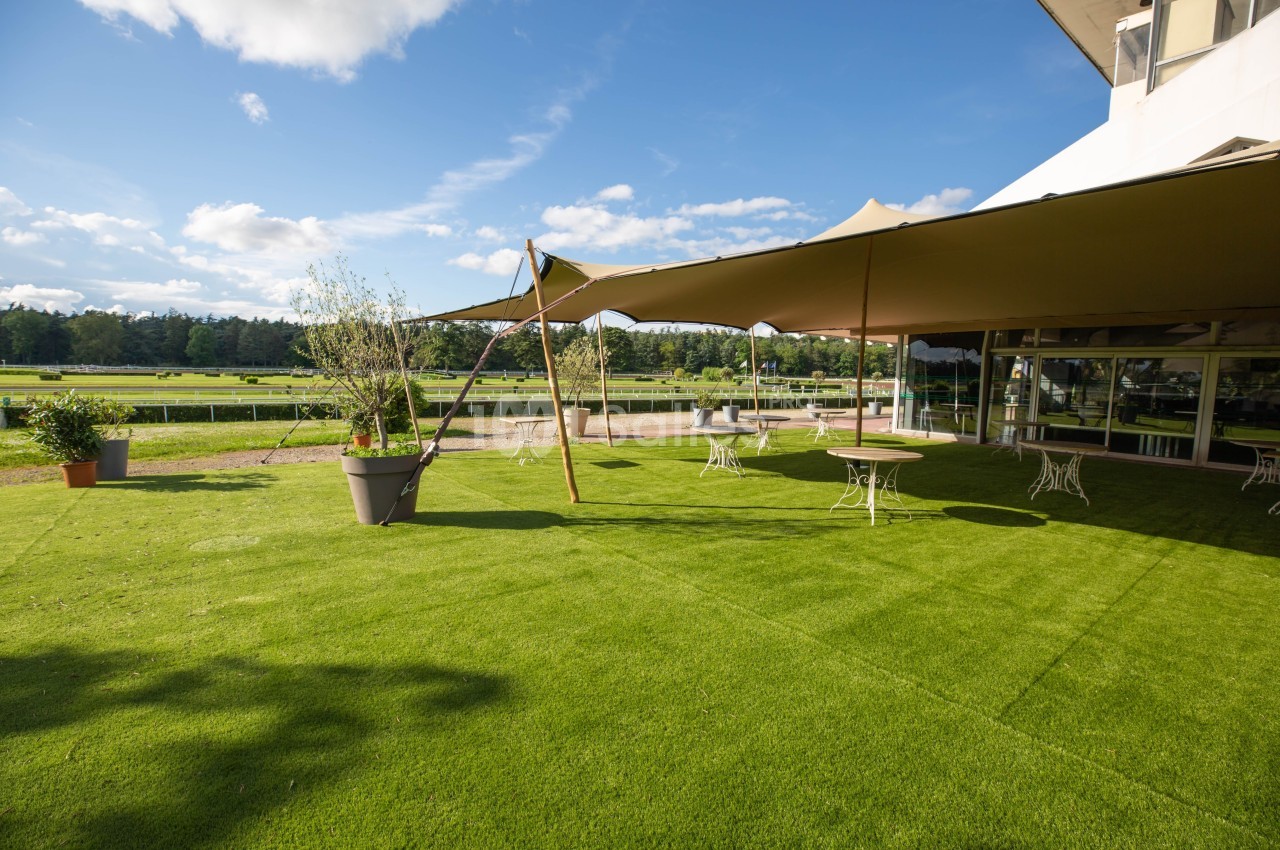 Espace extérieur avec pelouse, tables et chaises sous des toiles d'ombrage, entouré de verdure et d'un ciel dégagé.