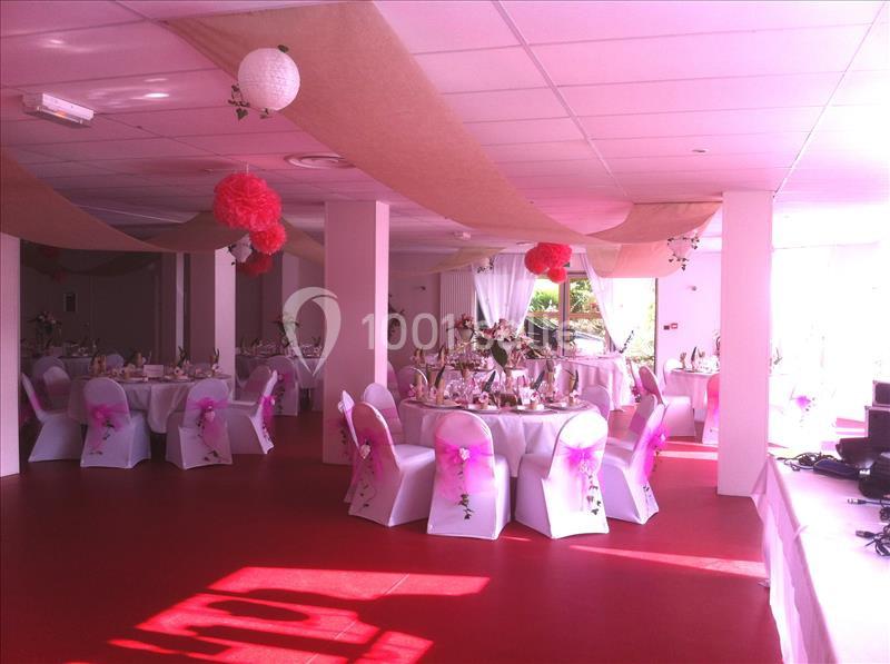 Salle de réception décorée avec des tables rondes, nappes blanches, chaises ornées de rubans roses et suspensions colorées.