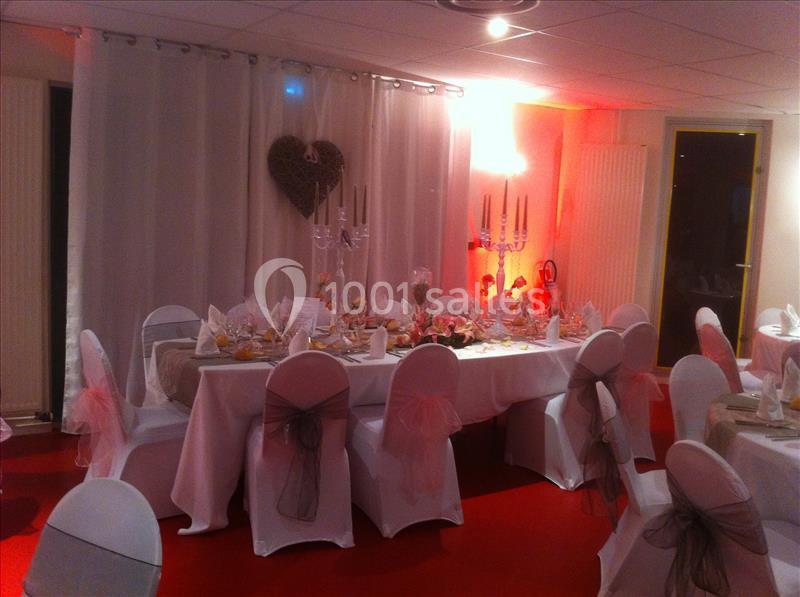 Salle décorée pour un événement, avec tables dressées, chaises couvertes et éclairage tamisé.