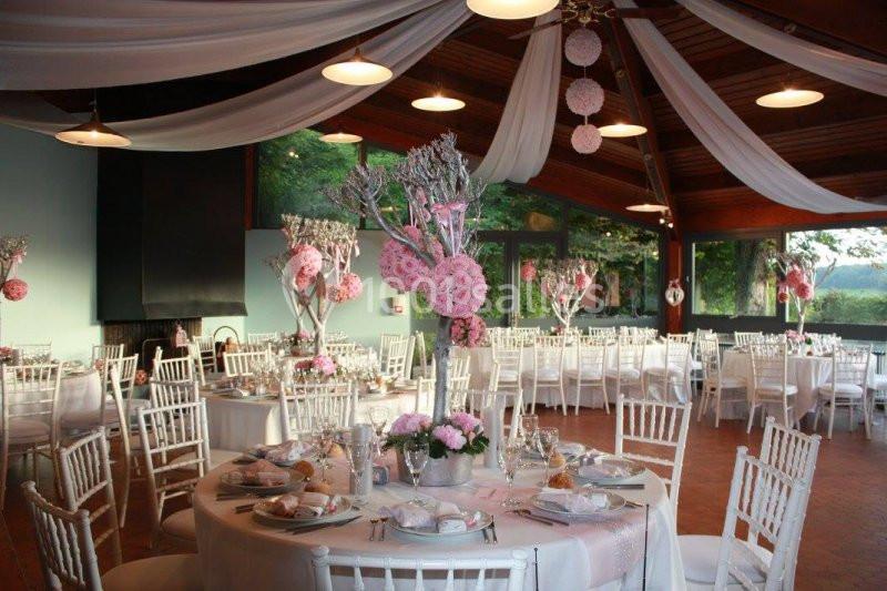 Salle de réception décorée avec des tables rondes, nappes blanches, chaises assorties et ornements floraux roses.