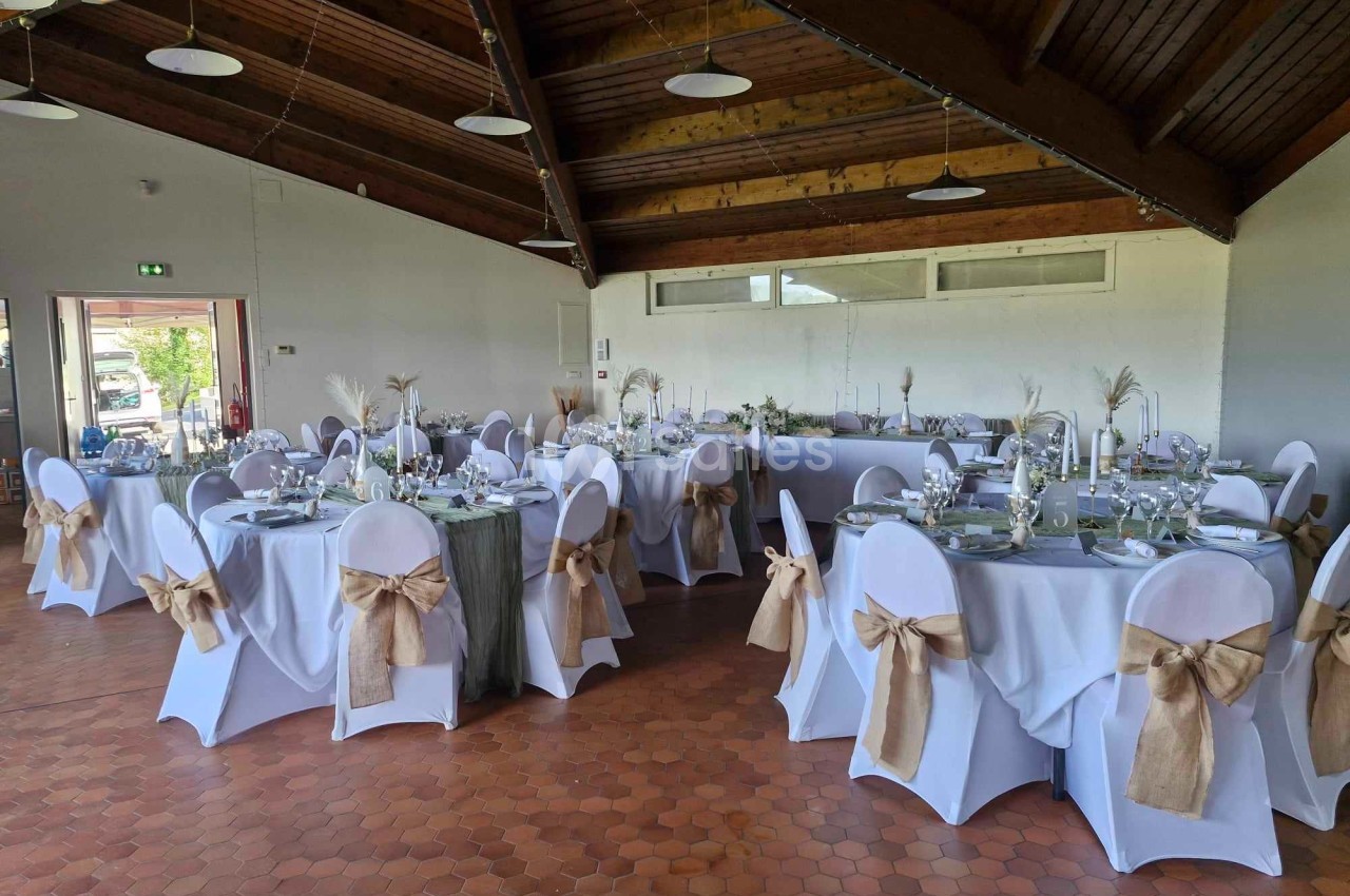 Salle de réception décorée avec des tables rondes, nappes blanches, chaises ornées de nœuds en toile de jute.