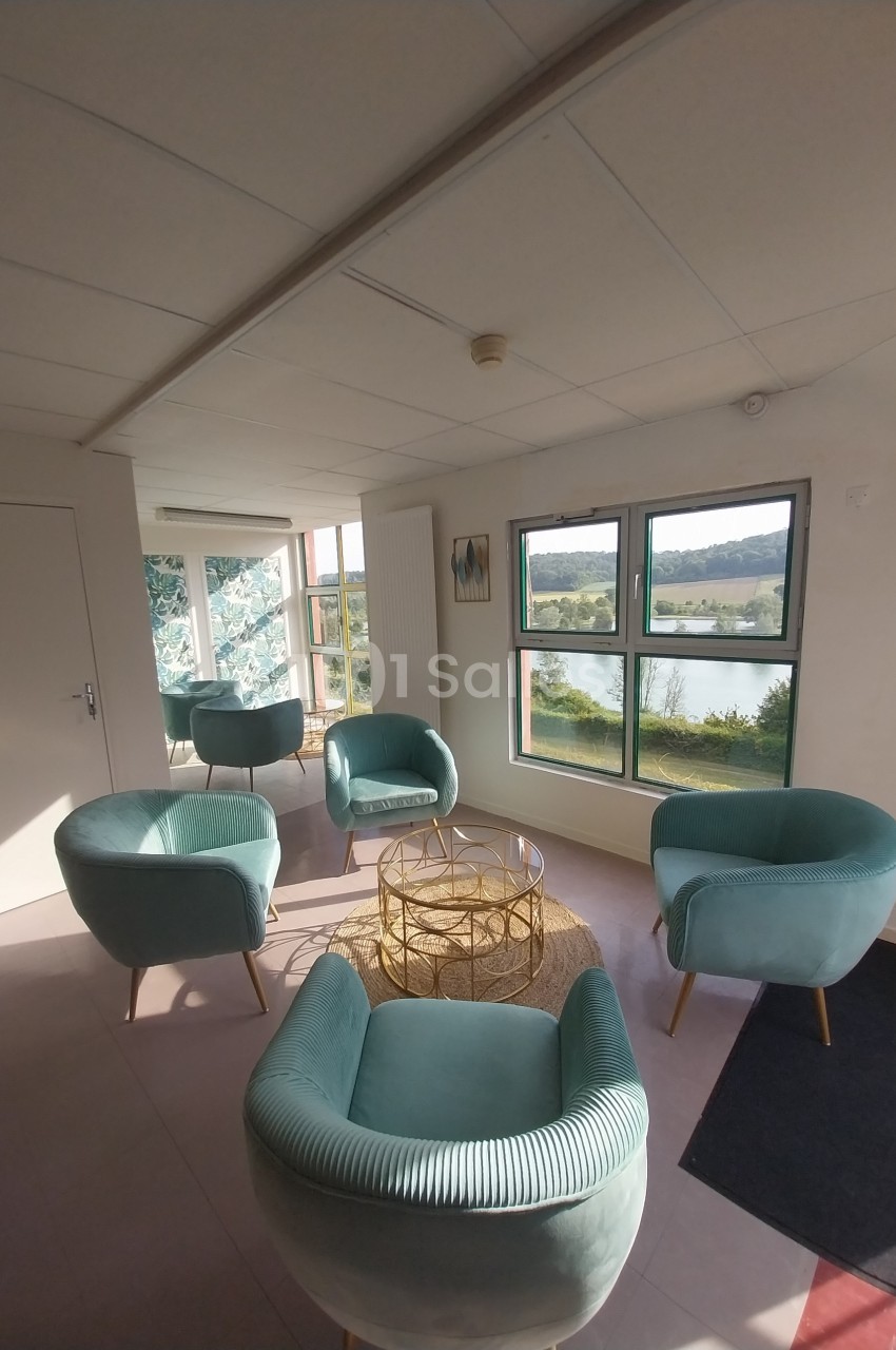 Espace lumineux avec fauteuils verts, table basse dorée et vue sur un paysage extérieur à travers de grandes fenêtres.