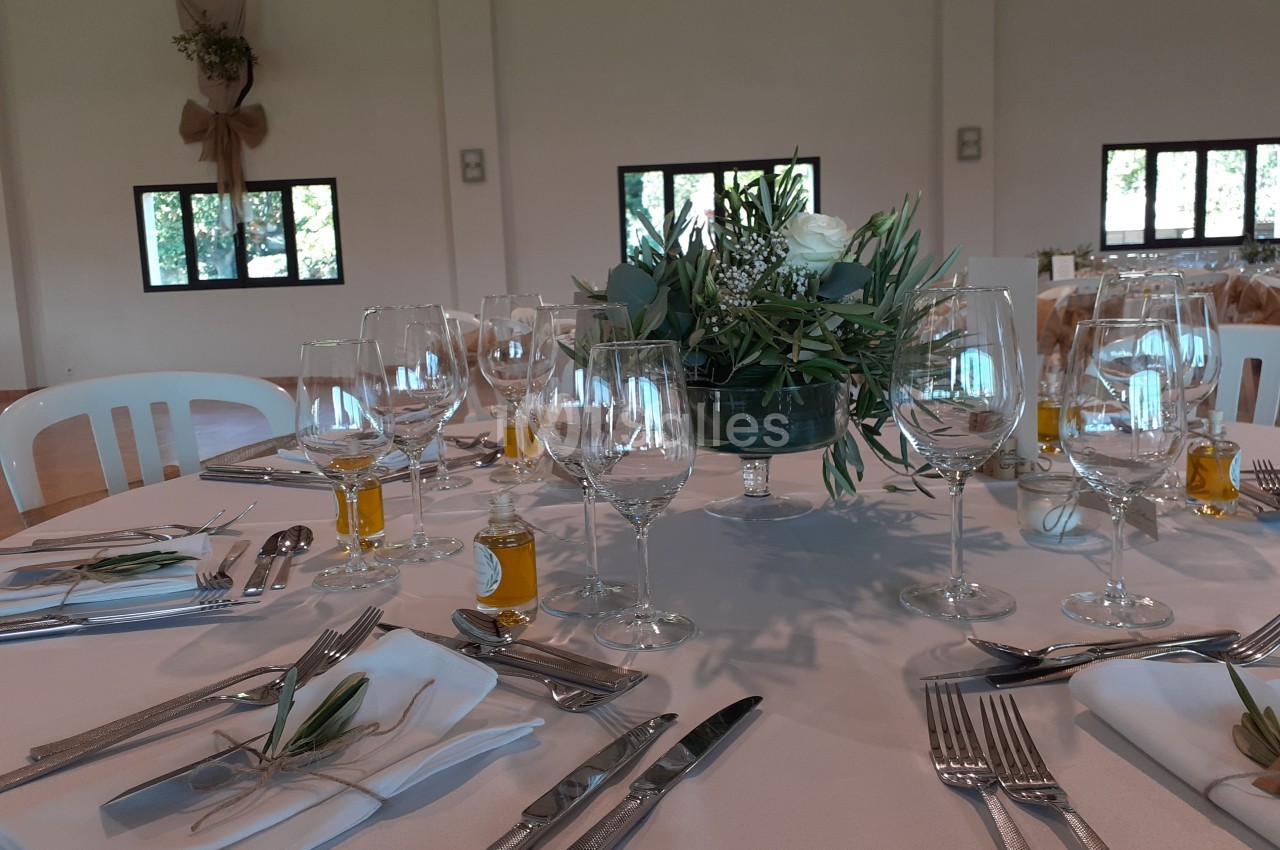 Table dressée pour un événement avec nappes blanches, couverts, verres, décorations végétales et petites bouteilles d'huile.