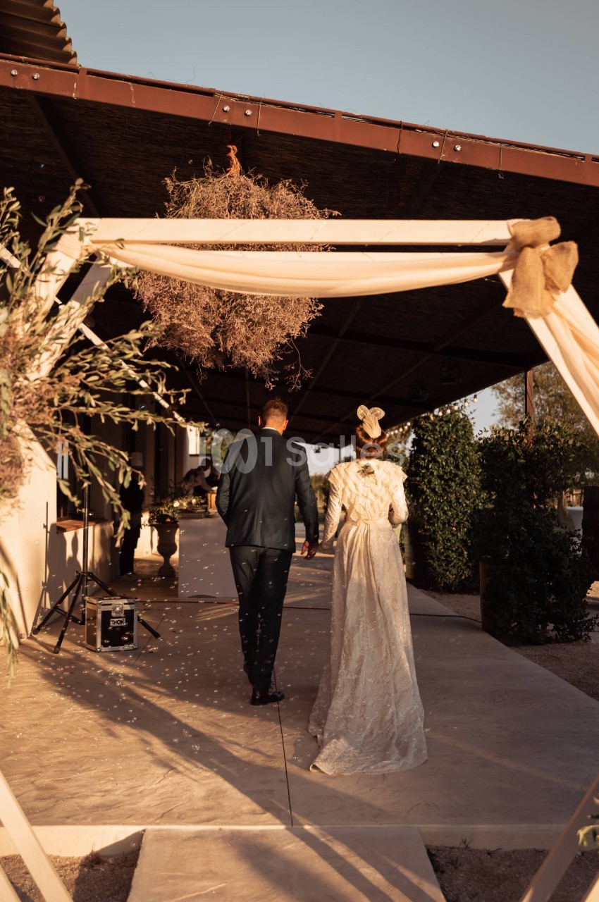 Un couple en tenue de mariage marche sous une arche décorée, éclairée par une lumière de fin de journée.