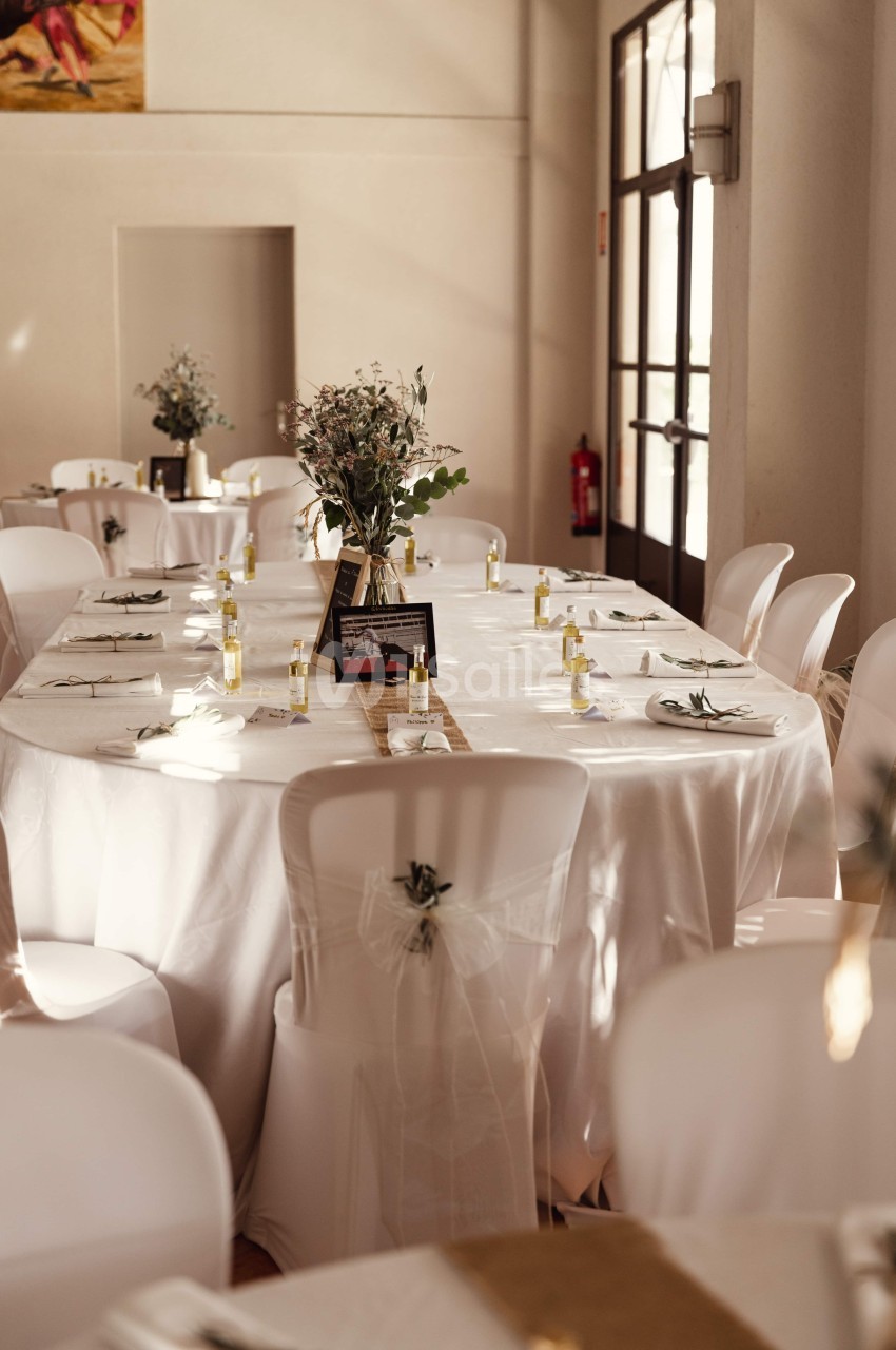 Salle de réception avec tables ovales dressées, nappes blanches, chaises décorées et centres de table floraux.