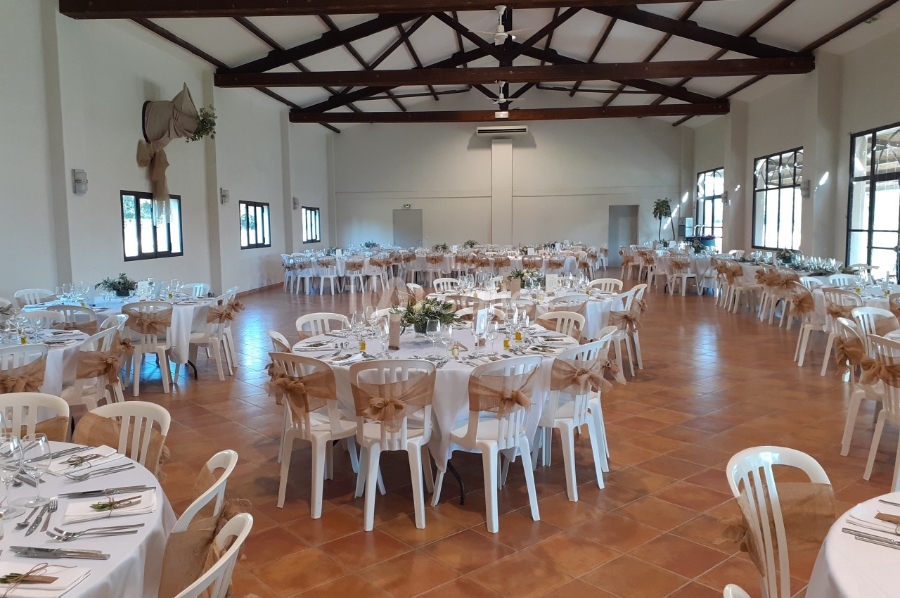 Salle de réception lumineuse avec tables rondes dressées, chaises blanches décorées de nœuds en toile de jute.
