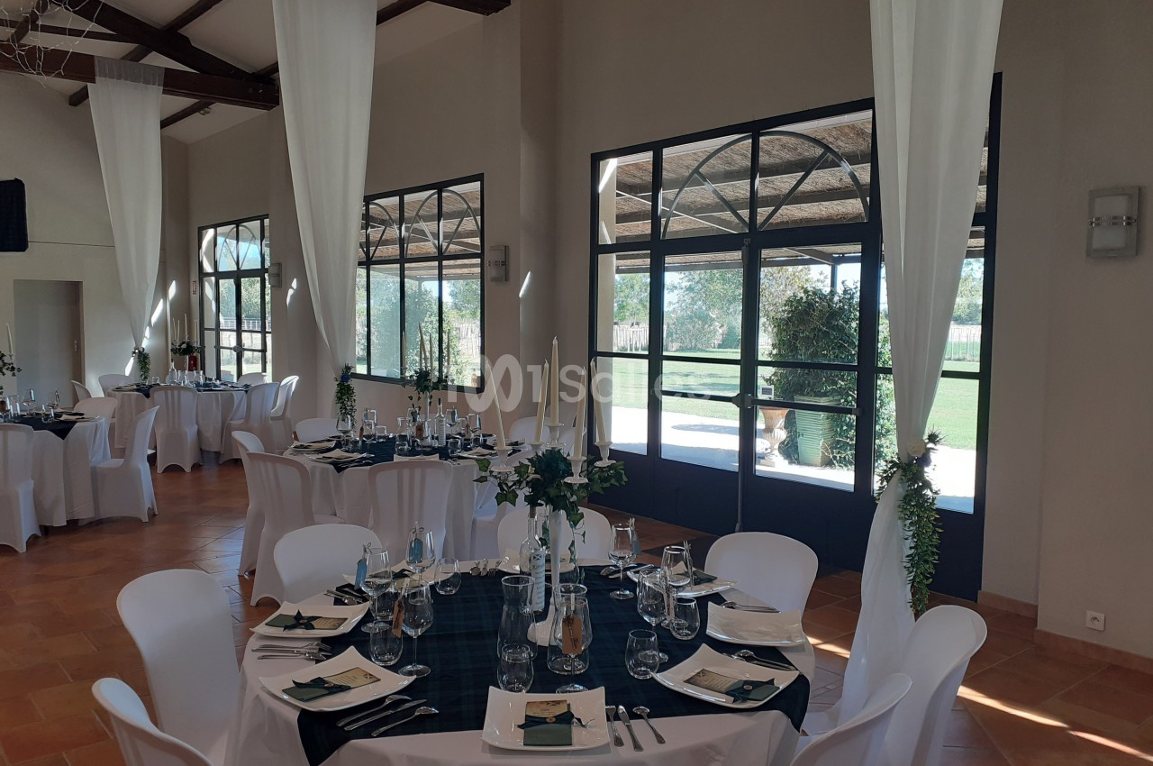 Salle de réception lumineuse avec tables dressées, nappes sombres, chaises blanches et grandes baies vitrées donnant sur un…