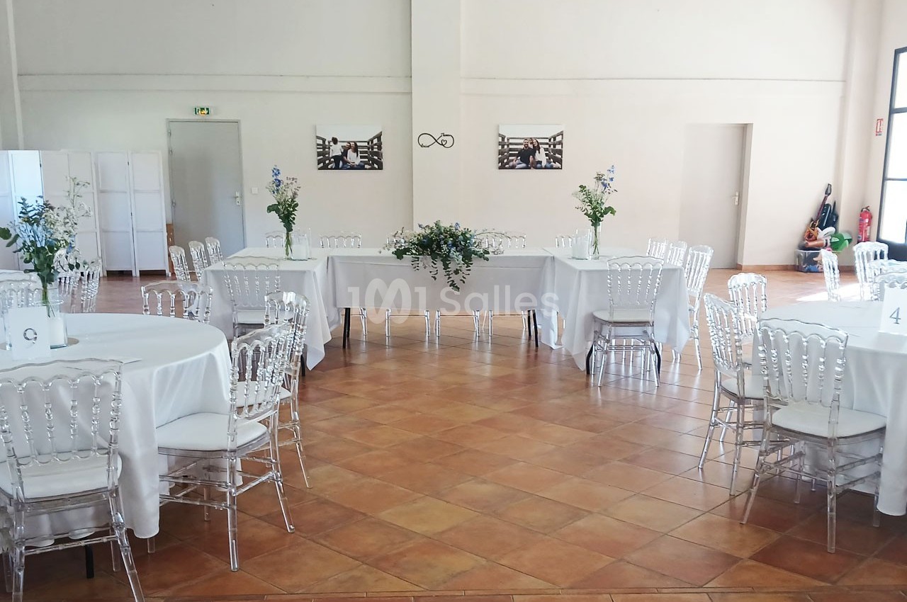Salle de réception lumineuse avec tables rondes dressées de nappes blanches, chaises transparentes et décorations florales.