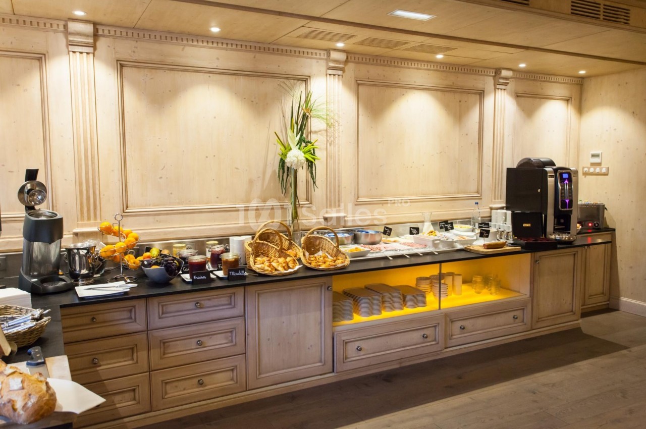 Buffet de petit-déjeuner avec viennoiseries, fruits, confitures, charcuterie et machine à café dans un décor en bois clair.