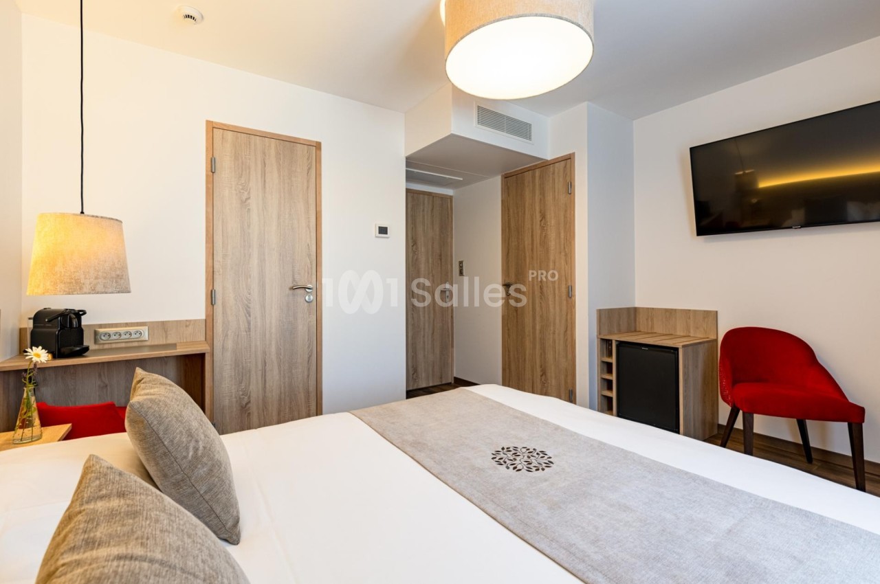 Chambre d'hôtel moderne avec lit double, télévision murale, bureau, chaise rouge et éclairage chaleureux.