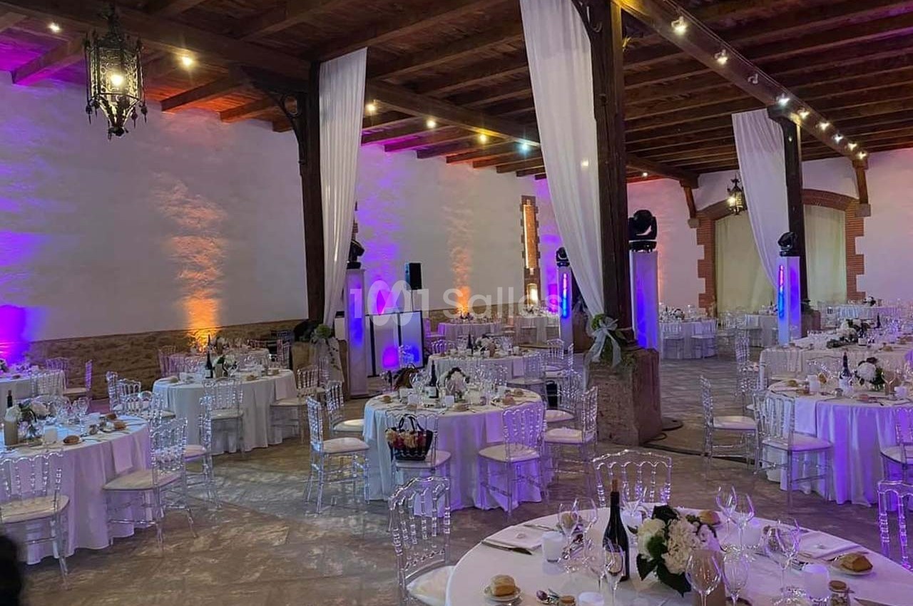 Salle de réception élégante avec tables rondes dressées, chaises transparentes et éclairage tamisé.