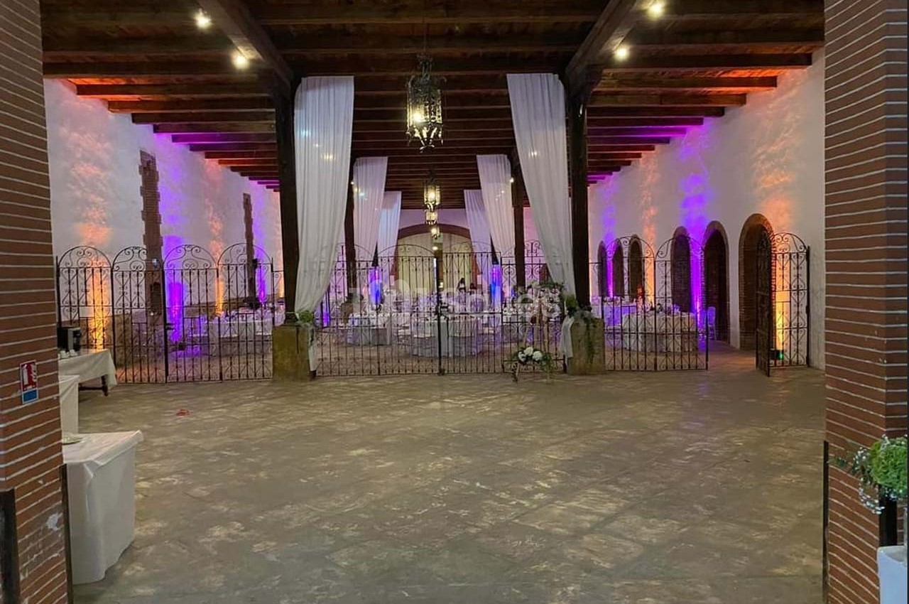 Salle de réception avec tables dressées, éclairage coloré et rideaux blancs suspendus sous un plafond en bois.