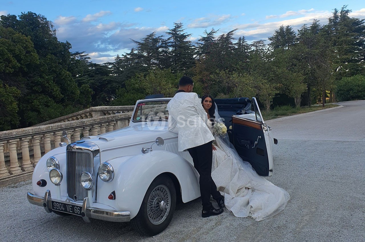 Un couple en tenue de mariage pose près d'une voiture ancienne blanche, dans un cadre extérieur arboré.