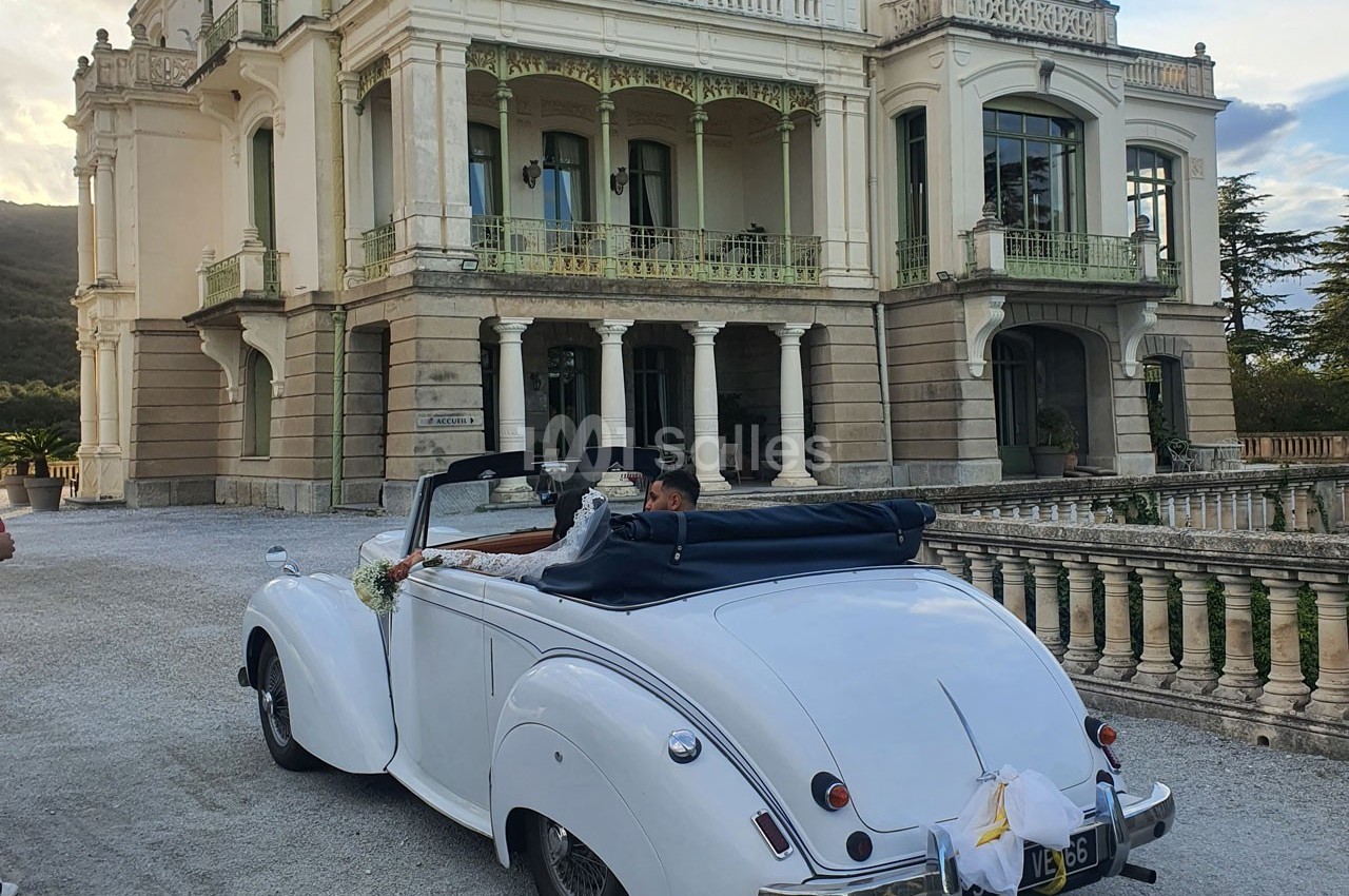 Une voiture ancienne décapotable blanche devant une grande bâtisse élégante, avec un couple à l'intérieur.