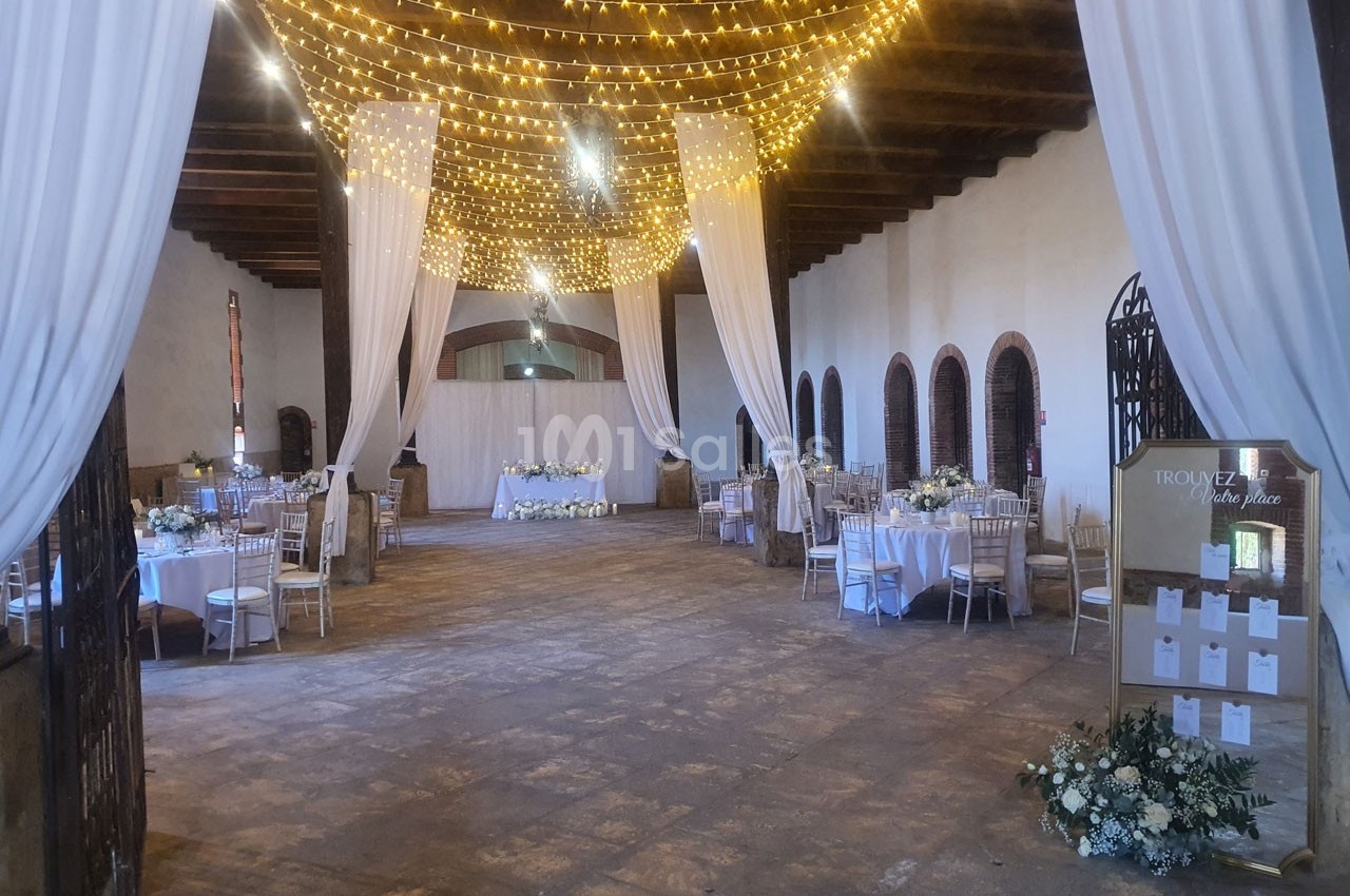 Grande salle de réception décorée avec des guirlandes lumineuses, des rideaux blancs et des tables élégamment dressées.