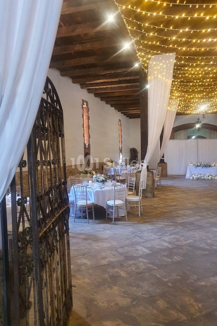 Salle de réception décorée avec des guirlandes lumineuses, des tables dressées et des rideaux blancs.