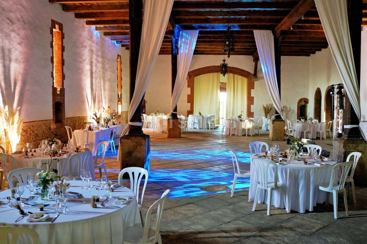 Salle de réception spacieuse avec tables rondes dressées, éclairage coloré et rideaux blancs suspendus.