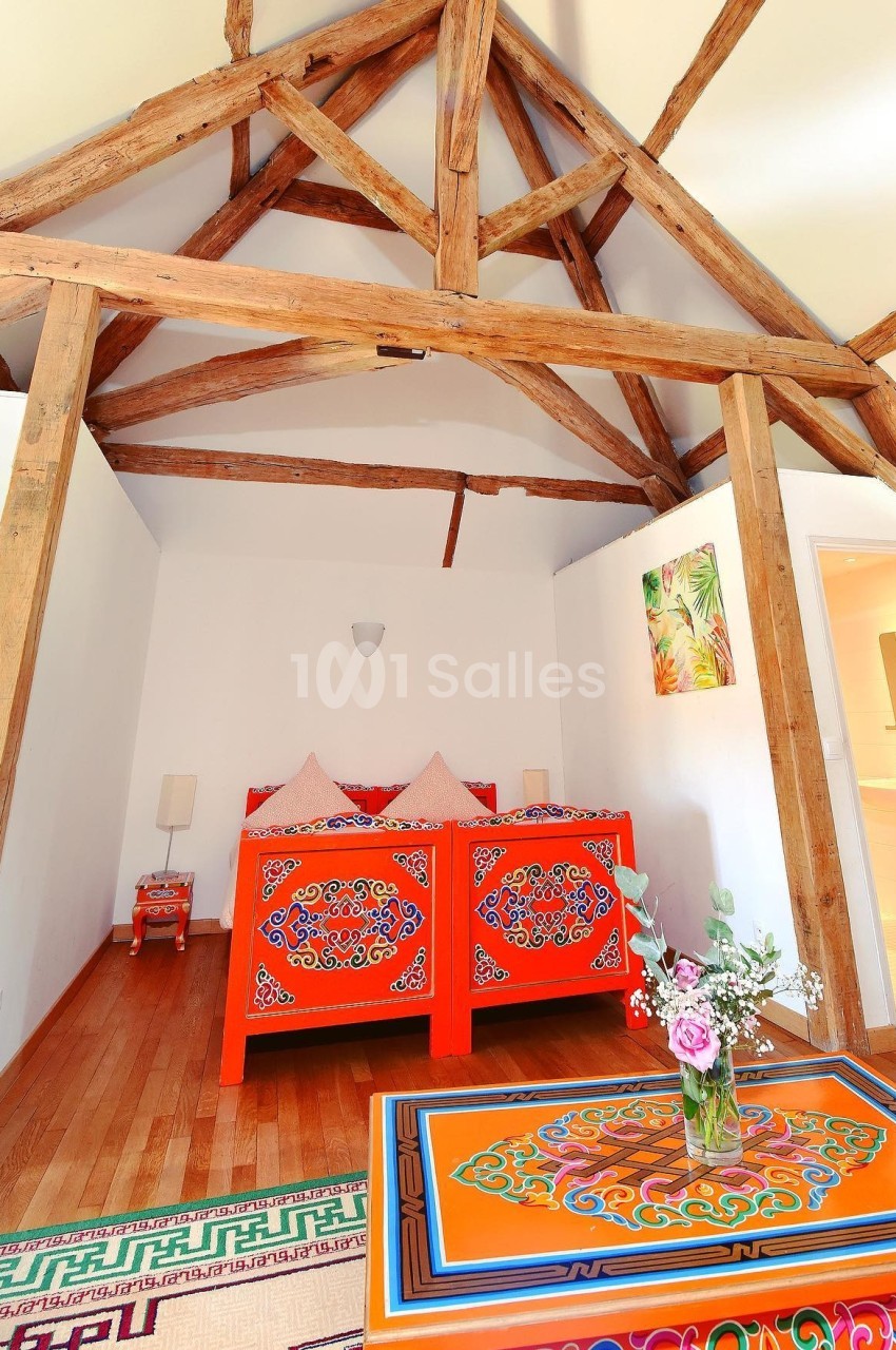 Chambre lumineuse avec lit en bois rouge sculpté, plafond à poutres apparentes et table décorée de motifs colorés.