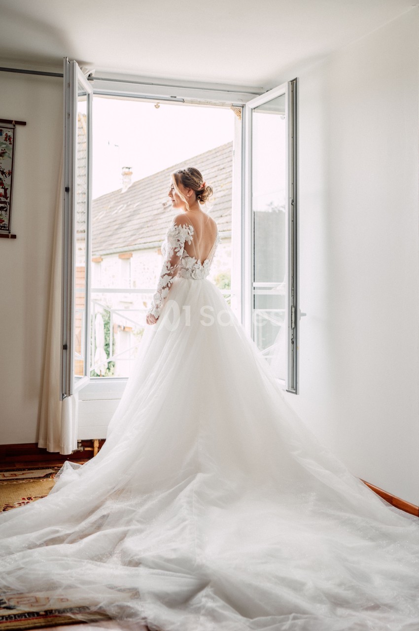 Femme en robe de mariée blanche, vue de dos, regardant par une fenêtre ouverte dans une pièce lumineuse.