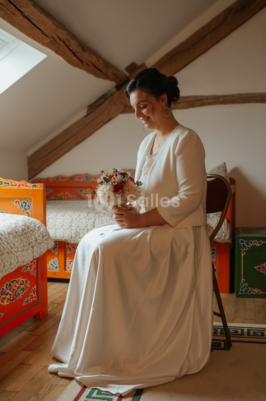 Femme en robe blanche assise sur une chaise, tenant un bouquet de fleurs, dans une pièce au décor traditionnel coloré.