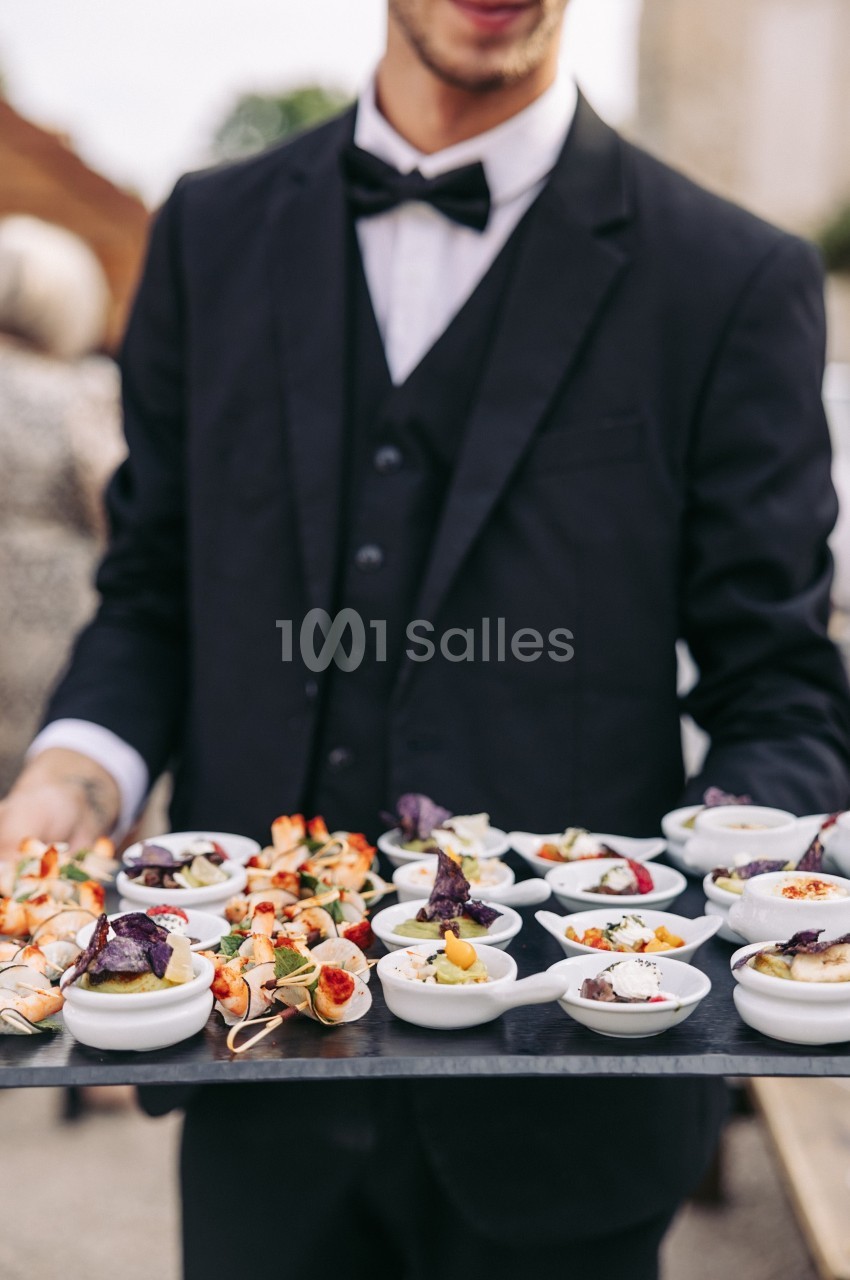 Serveur en costume portant un plateau de petites assiettes garnies d'amuse-bouches variés lors d'un événement.
