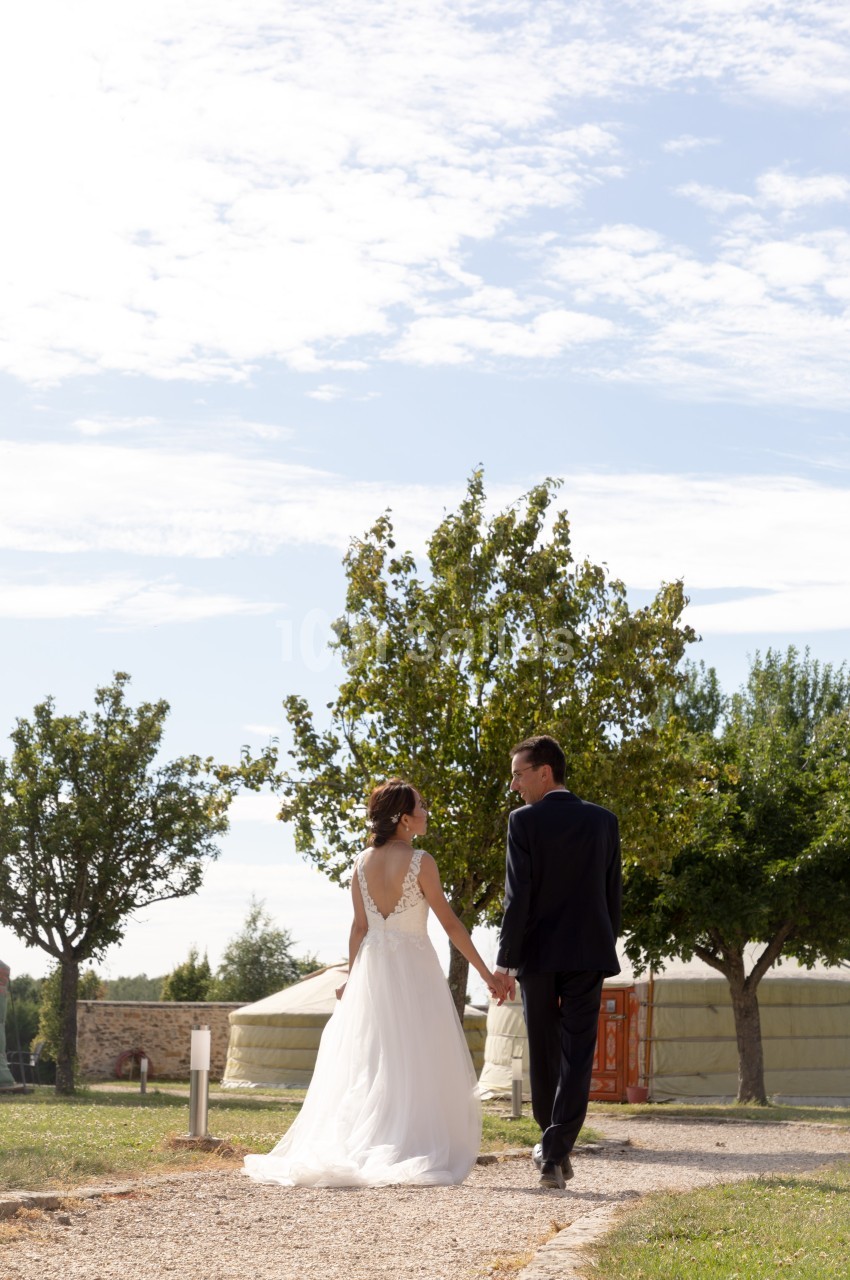 Un couple en tenue de mariage marche main dans la main sur un chemin, entouré d'arbres et de yourtes sous un ciel dégagé.