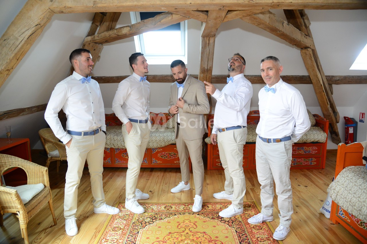 Cinq hommes en tenue élégante, dont un en costume beige, posent dans une pièce avec des poutres apparentes.