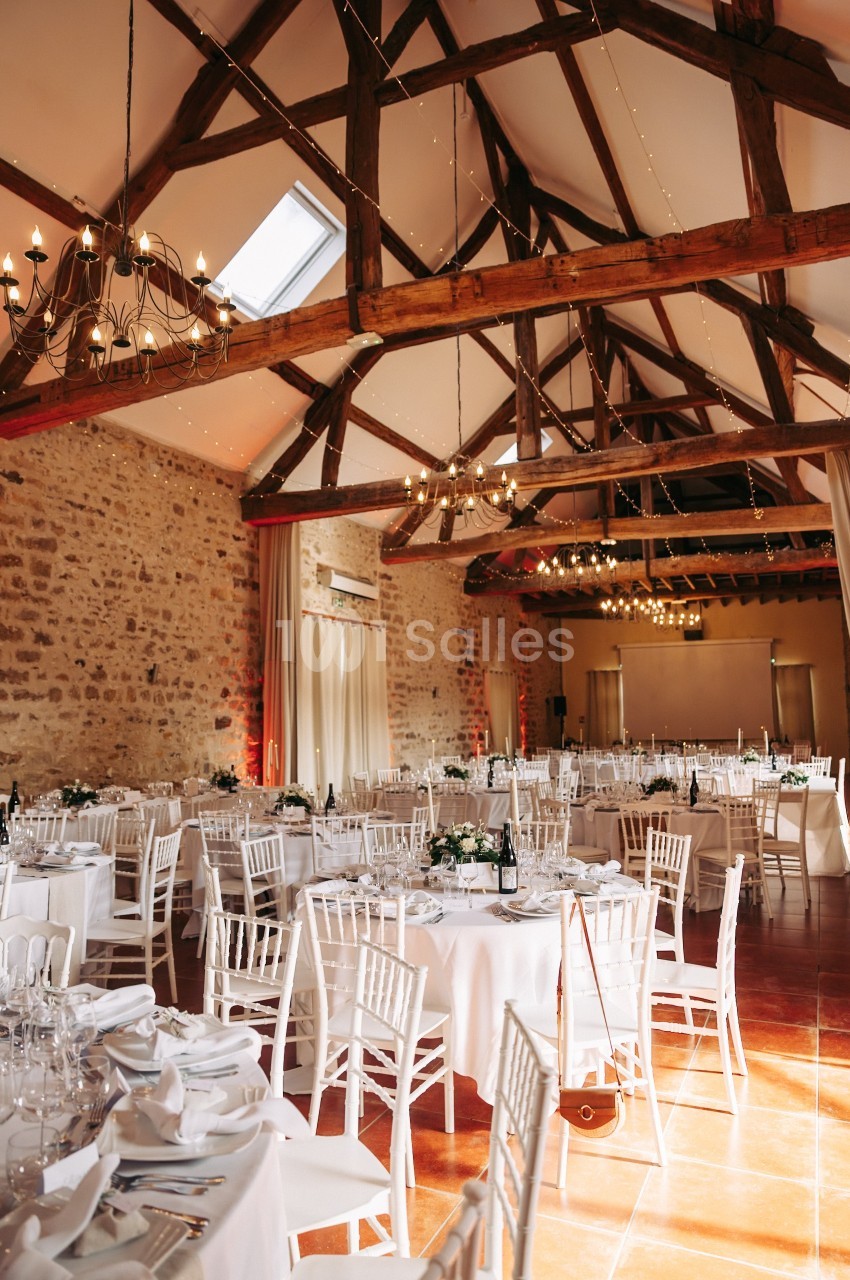 Salle de réception lumineuse avec murs en pierre, poutres apparentes et tables rondes décorées pour un événement.