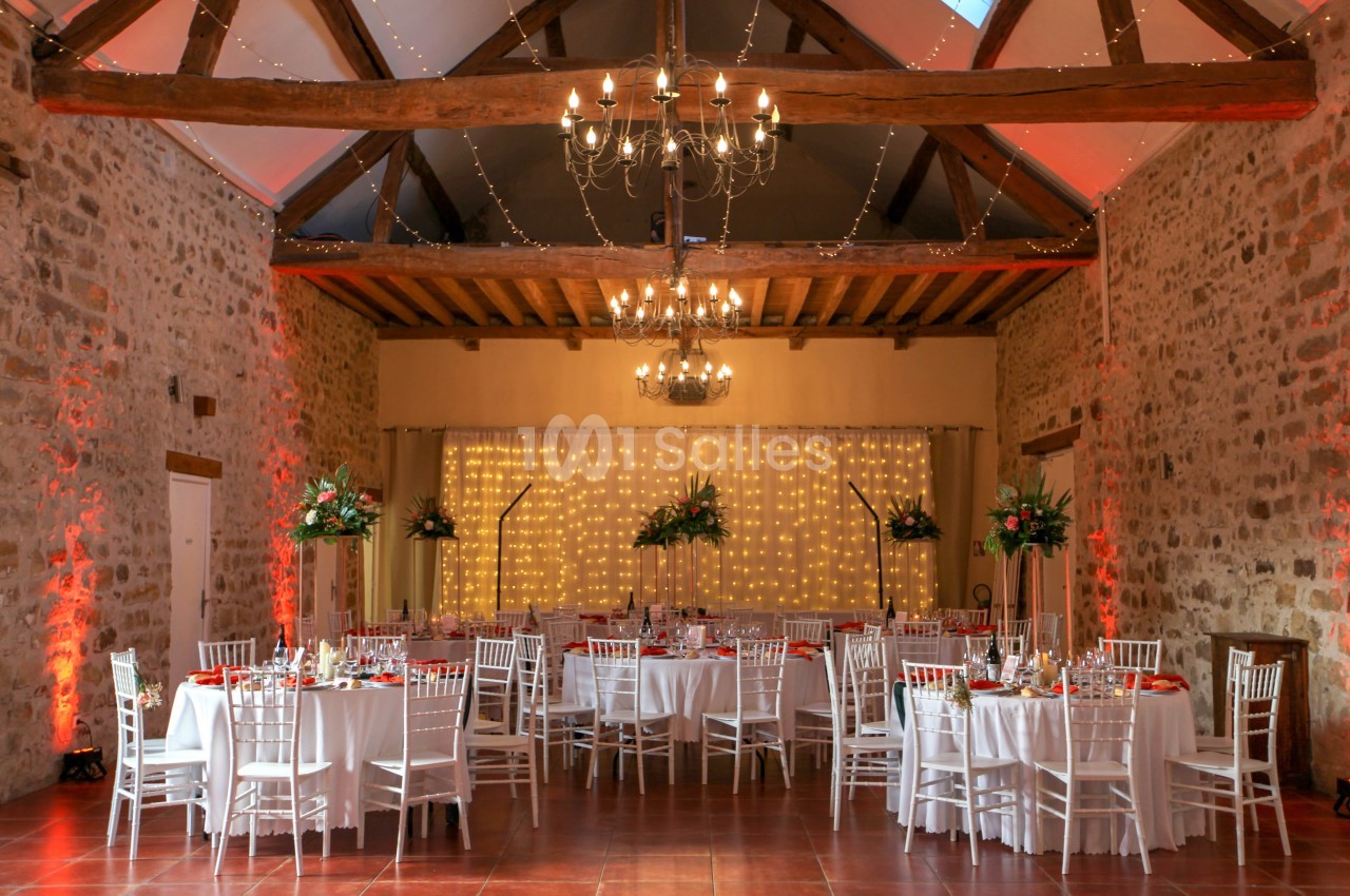 Salle de réception en pierre avec tables rondes décorées, chaises blanches et guirlandes lumineuses suspendues.