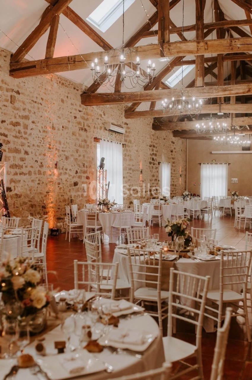 Salle de réception avec murs en pierre, poutres apparentes et tables rondes décorées pour un événement.