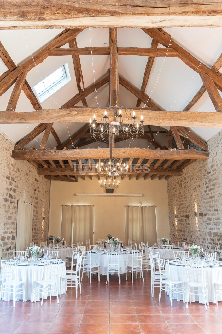Salle de réception lumineuse avec poutres apparentes, tables rondes dressées et chaises blanches.