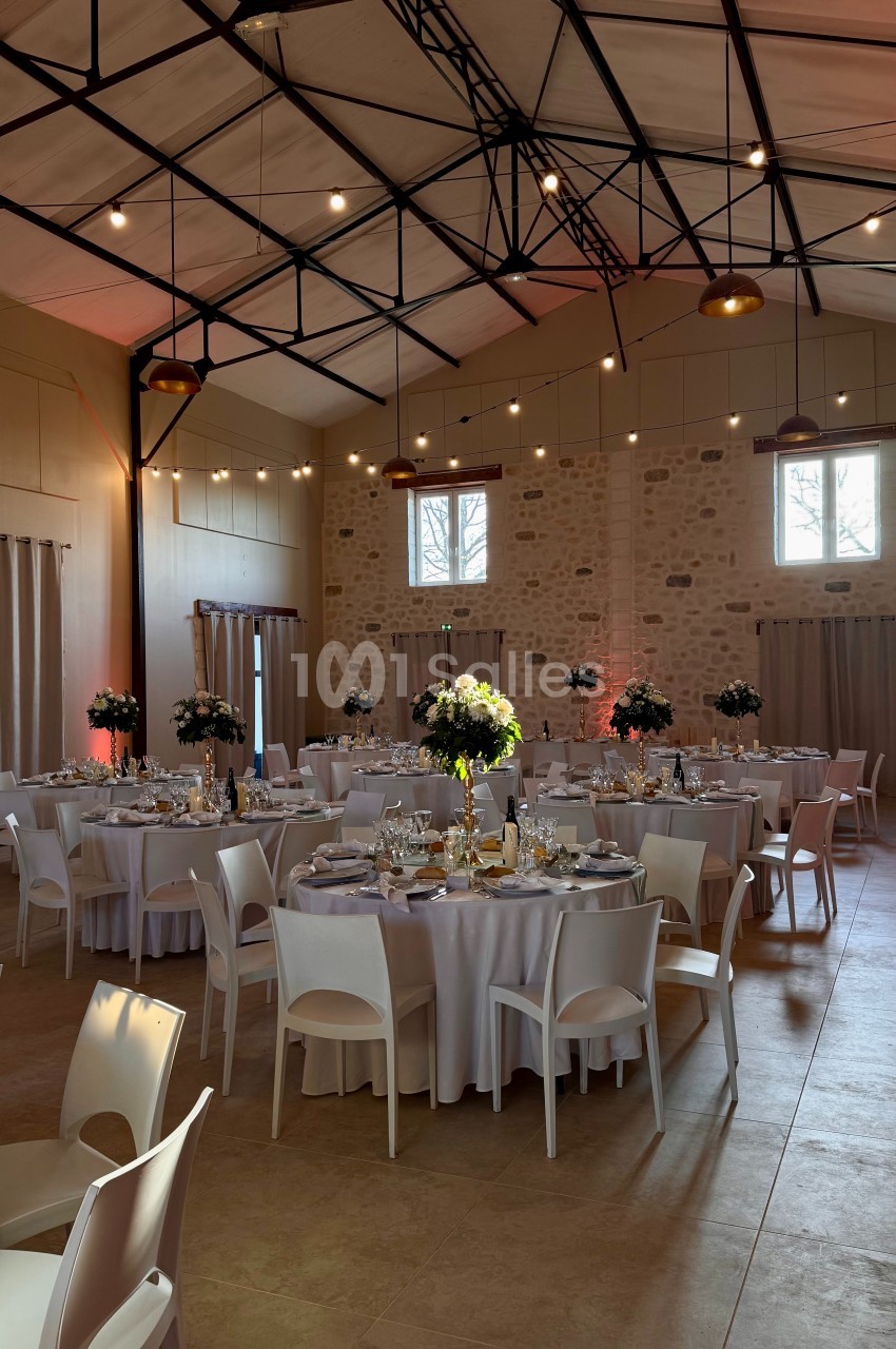 Salle de réception décorée pour un événement, avec tables rondes dressées, nappes blanches et arrangements floraux.