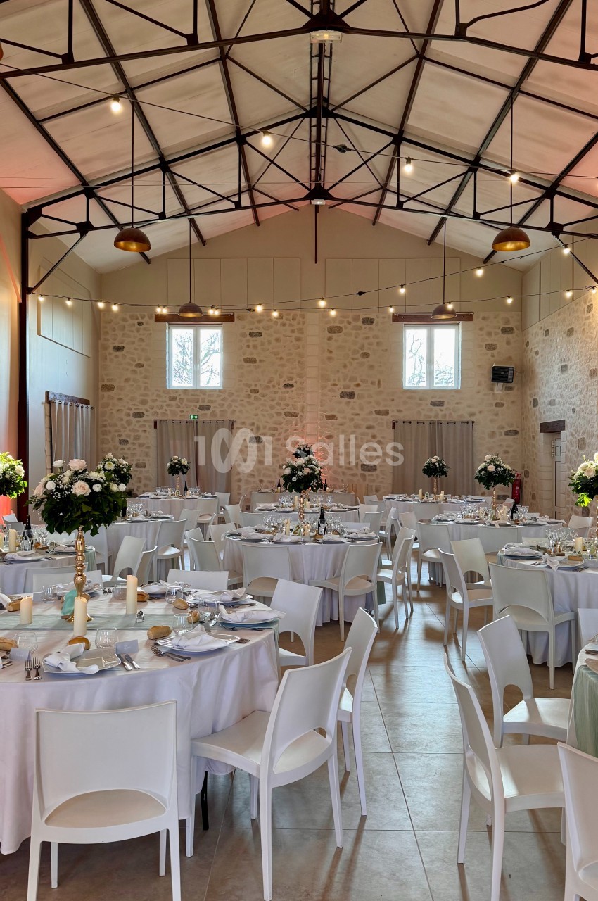 Salle de réception lumineuse avec tables rondes dressées, chaises blanches et décorations florales élégantes.