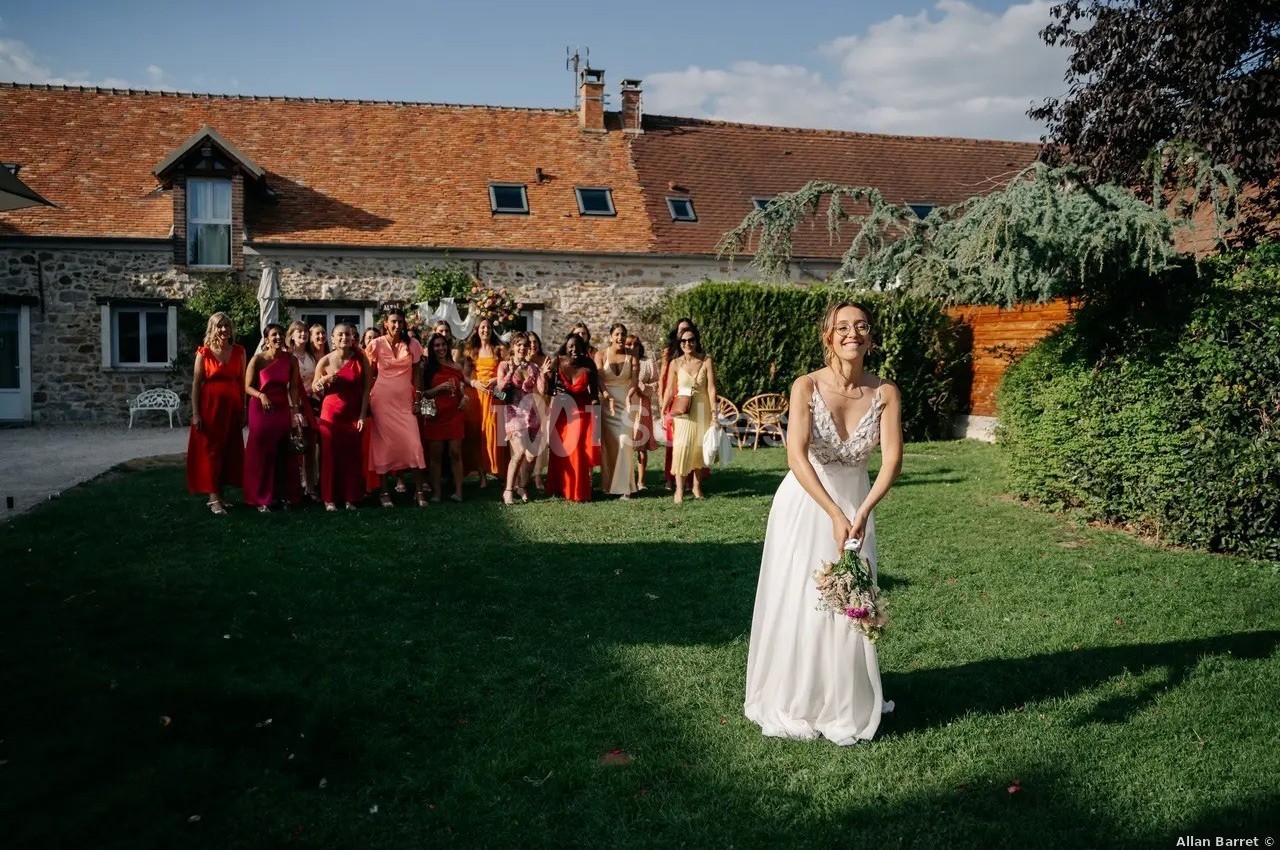 Une mariée en robe blanche s'apprête à lancer son bouquet devant un groupe d'invitées dans un jardin ensoleillé.