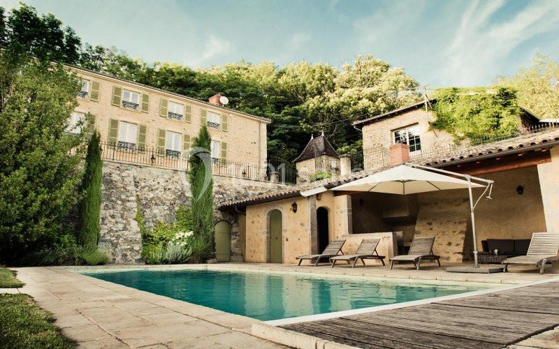 Maison en pierre avec piscine, terrasse aménagée et jardin arboré, située au pied d'une colline verdoyante.