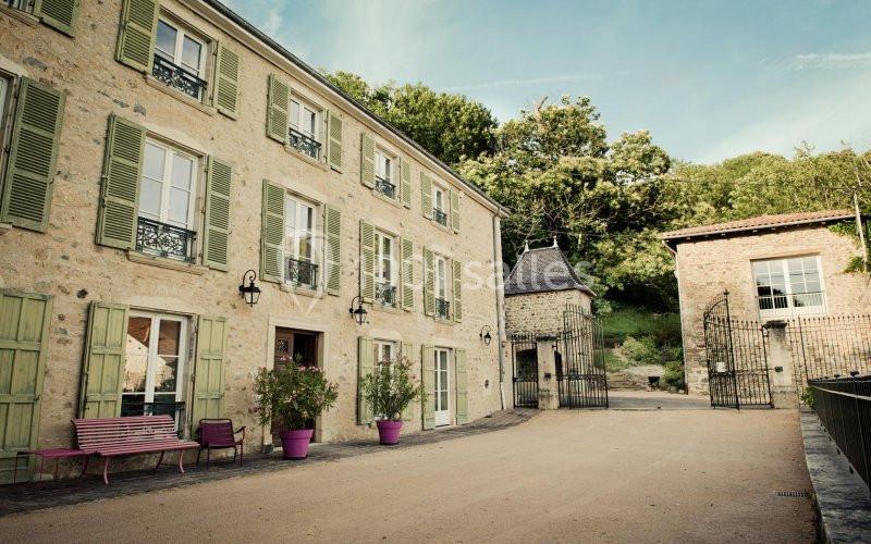 Façade d'un bâtiment en pierre avec volets verts, cour pavée, banc rose et végétation en arrière-plan.
