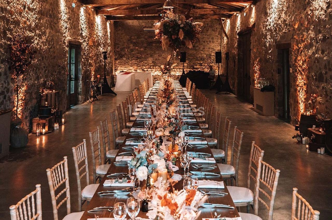 Salle en pierre avec une longue table décorée de fleurs et bougies, éclairée par une lumière chaleureuse.