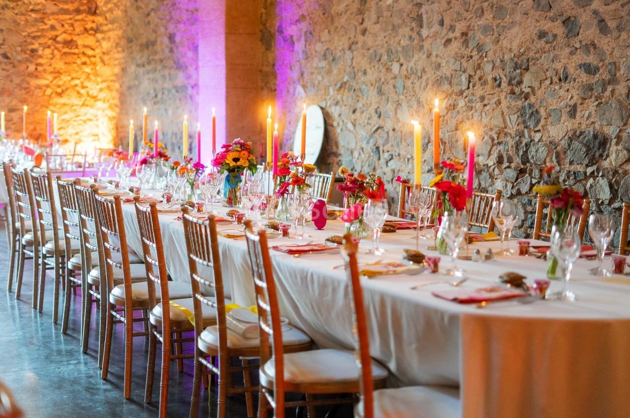Grande table décorée pour un événement, avec nappes blanches, chandeliers allumés et arrangements floraux colorés.