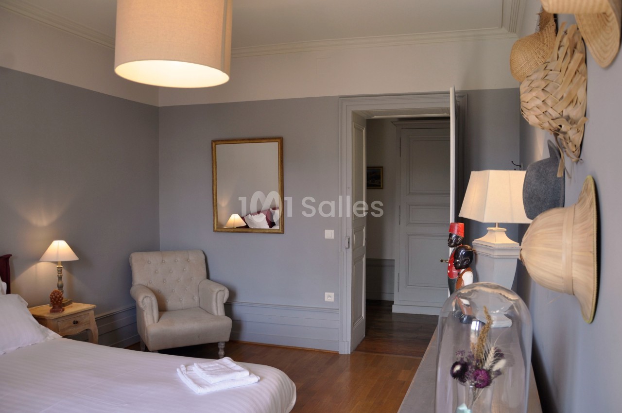 Chambre lumineuse avec murs gris, lit, fauteuil beige, miroir doré, lampes allumées et décoration murale en chapeaux.
