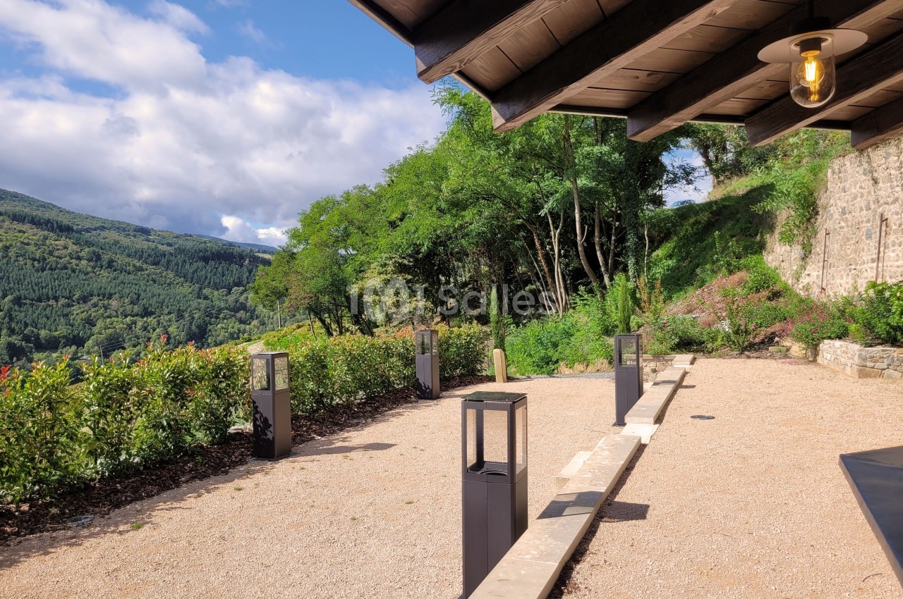 Terrasse en gravier avec des lanternes modernes, bordée de végétation et offrant une vue sur des collines verdoyantes.