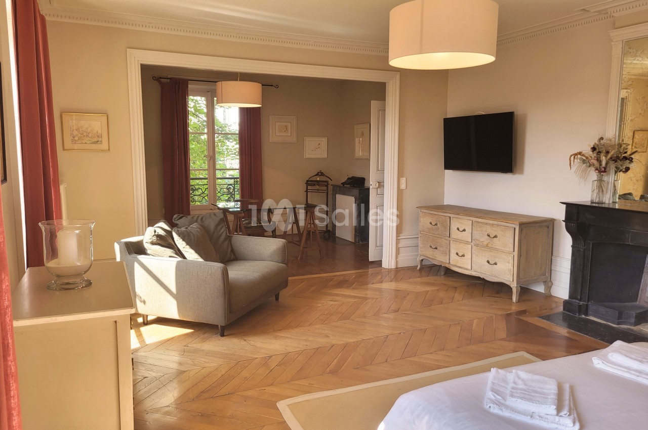 Salon lumineux avec parquet en bois, canapé, bureau, cheminée, télévision murale et grandes fenêtres avec rideaux rouges.