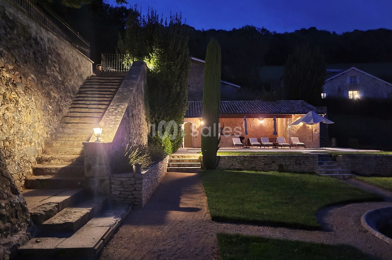 Cour extérieure éclairée de nuit avec escaliers en pierre, piscine, chaises longues et bâtiments en arrière-plan.