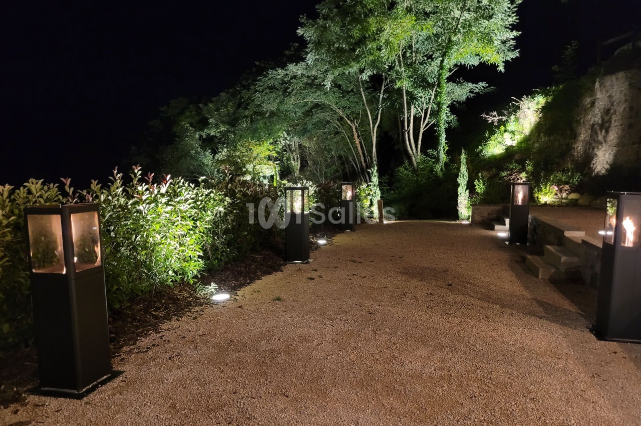 Allée extérieure éclairée la nuit par des lanternes modernes et des spots au sol, entourée de végétation.