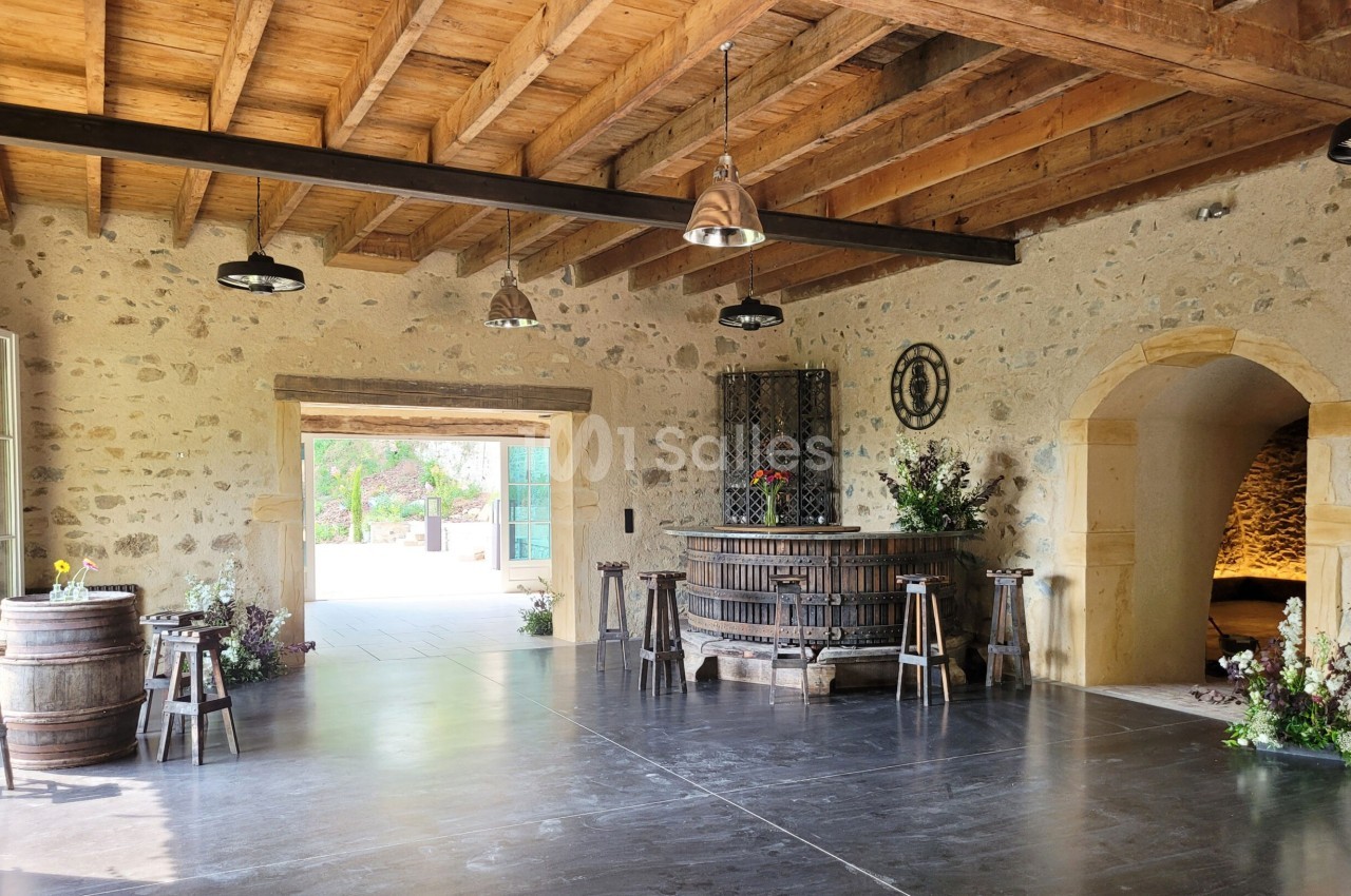 Salle en pierre avec un plafond en bois, un ancien pressoir au centre, des tonneaux et une ouverture sur l'extérieur.