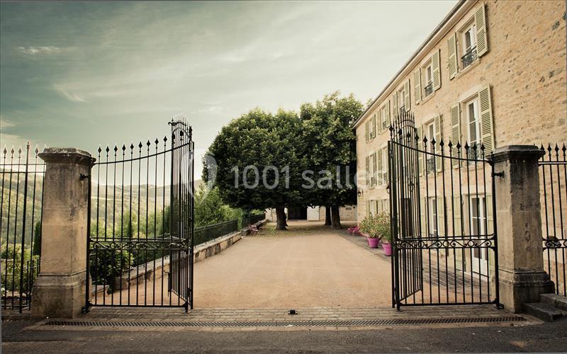 Entrée d'une propriété avec portail en fer forgé ouvert, allée bordée d'arbres et bâtiment en pierre à droite.